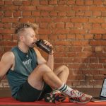 Athletic man sitting on exercise mat with water bottle, taking a break from workout, in a rustic indoor setting.