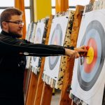 Man in glasses practicing archery indoors, pulling arrows from bullseye on target.